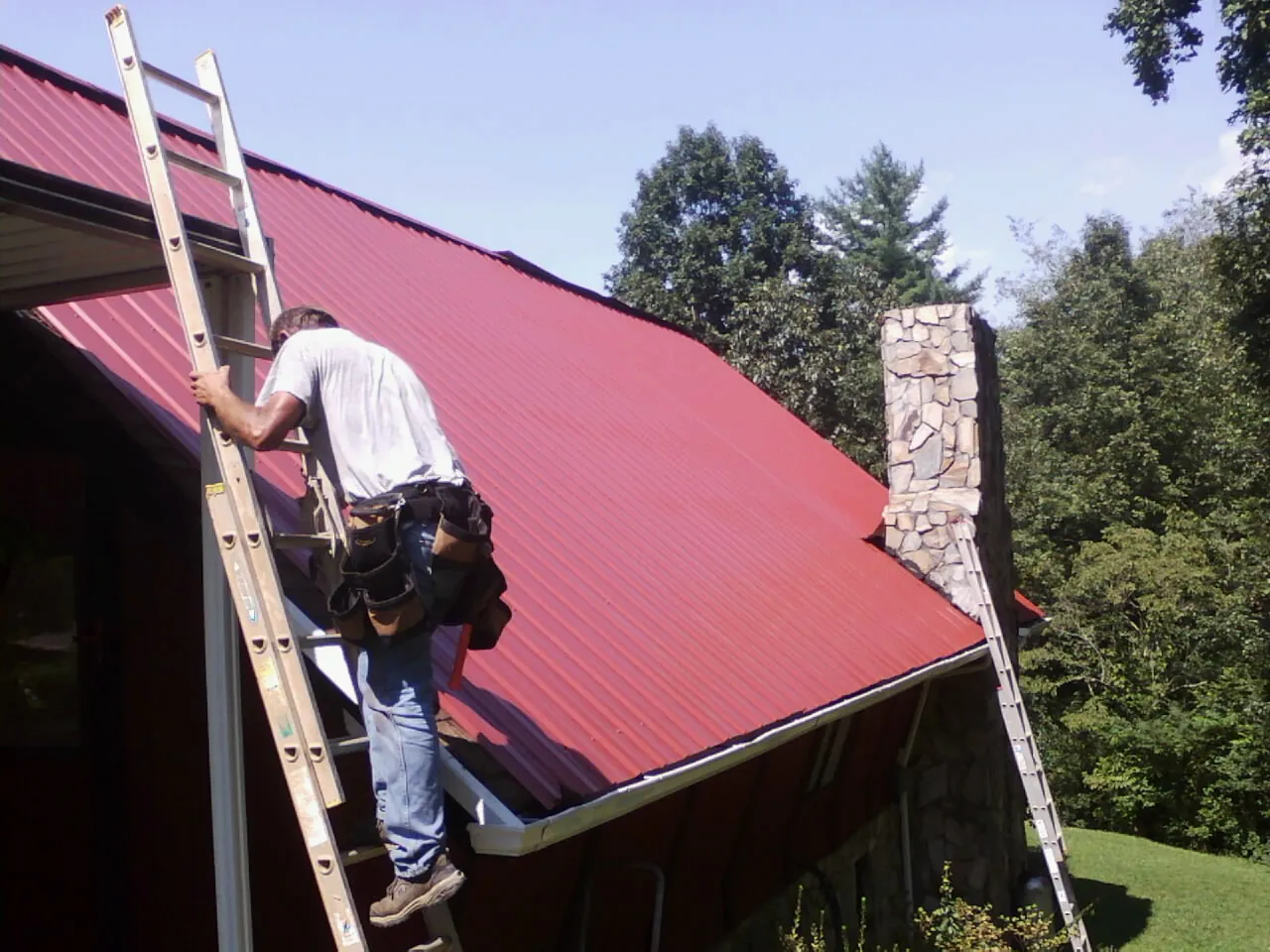 Roofer on a ladder beside a red metal roof and stone chimney — Soffit & Fascia Repair by Roofing Brothers in Jobstown